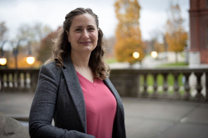 Photo of a woman wearing a pink shirt and gray blazer smiling and standing outside. From the colors of the trees it looks to be fall